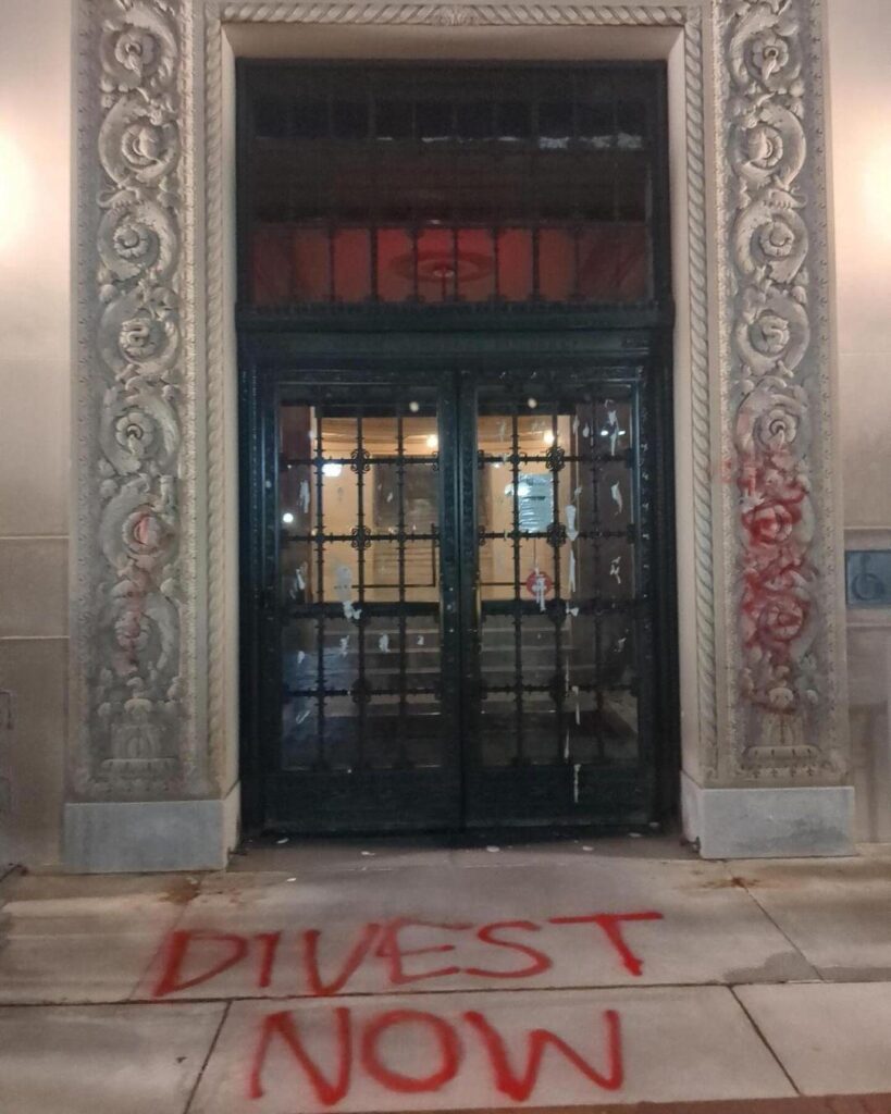 un tag « Désinvestissez maintenant » sur le sol devant un bâtiment, de la peinture rouge sur la façade, et de nombreuses traces de peinture blanche sur une porte vitrée
