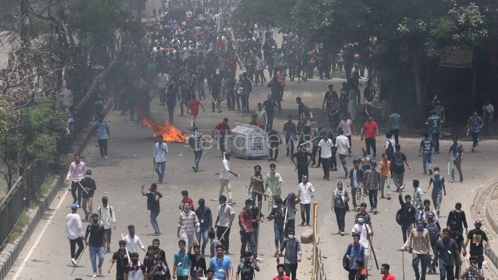 Une foule de manifestants affronte la police dans les manifestations du 18 juillet à Dacca. Nombre d'entre eux sont armés de bâtons ou masqués, un feu se trouve au milieu de la foule.
